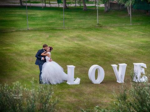 Bride and groom embracing near LOVE letters on green lawn.