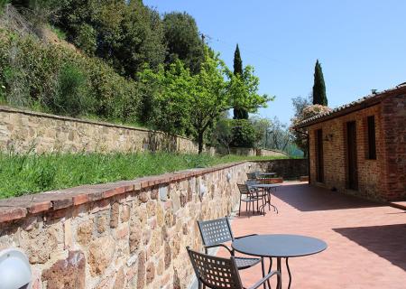 Terrazza soleggiata con tavoli, mura in pietra e alberi verdi.