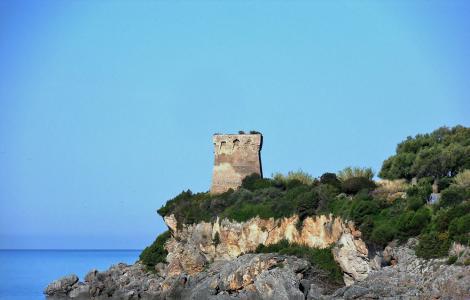 Torre antica su scogliera, mare calmo, cielo azzurro, natura selvaggia.