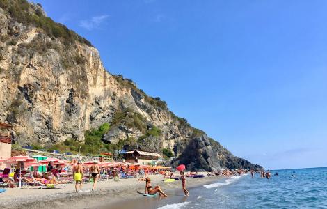 Spiaggia affollata, mare blu, scogliere imponenti, sole splendente, estate vivace.