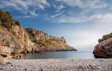 Spiaggia di ciottoli, mare calmo, scogliere maestose, cielo azzurro, atmosfera serena.