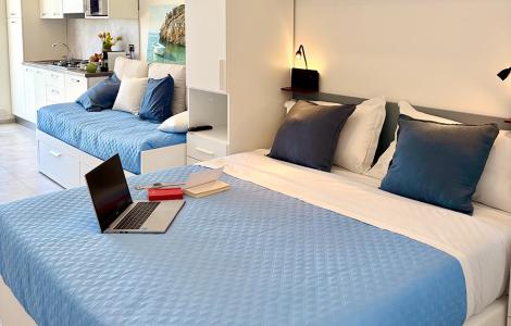 Camera luminosa con letto, divano e laptop su copriletto azzurro.