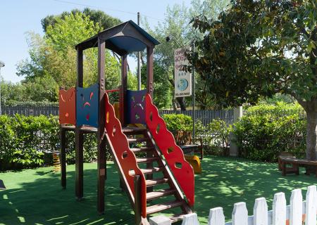 Parco giochi colorato con scivolo, alberi verdi e sole splendente.