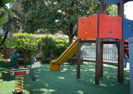 Parco giochi soleggiato con scivolo giallo e giochi colorati sotto un grande albero.