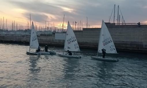 Tre barche a vela navigano al tramonto in un porto.