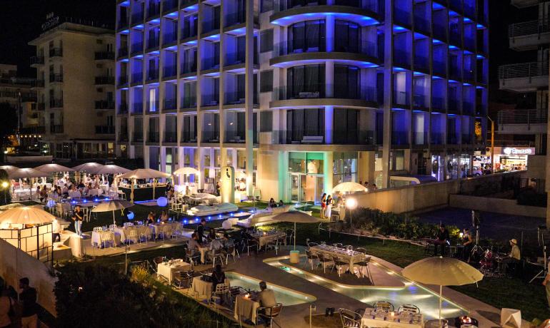 Hotel elegante illuminato di blu, atmosfera vivace con tavoli all'aperto e musica.