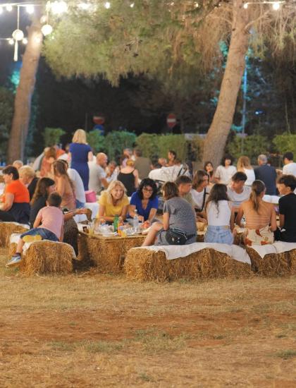 Outdoor party with straw tables, lights, and people socializing.