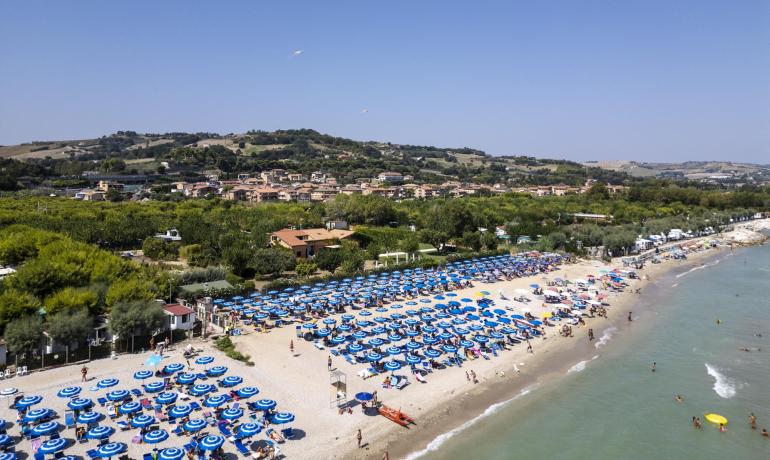 Spiaggia affollata con ombrelloni blu e colline sullo sfondo.