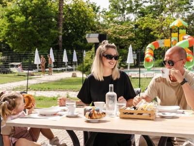 Famiglia felice fa colazione all'aperto, vicino a una piscina e giochi colorati.