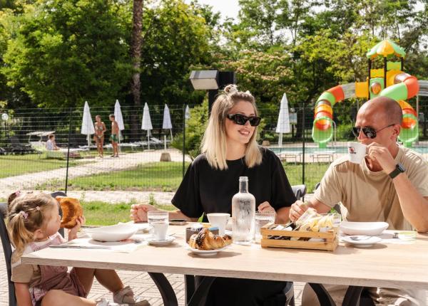 Famiglia felice fa colazione all'aperto, vicino a una piscina e giochi colorati.